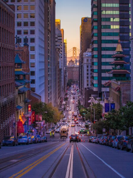 A steep San Francisco street view with cars, buildings, and the distant Bay Bridge at twilight.