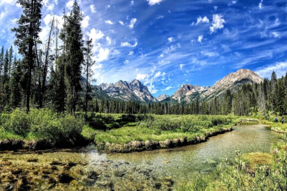 A picturesque view of Idaho's Sawtooth Mountains, featuring a clear stream, lush greenery, forest, and a partly cloudy sky.