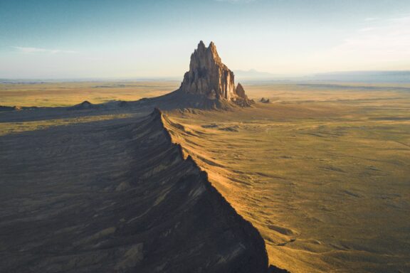 Daylight aerial view of Shiprock, a notable rock formation in New Mexico, amidst a flat desert landscape.