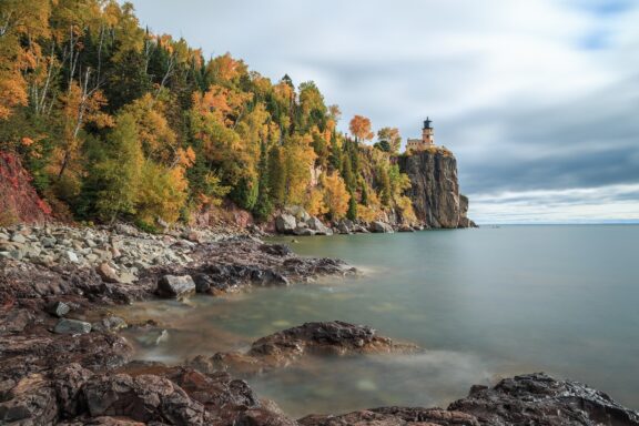 A lighthouse atop a cliff with autumn-colored trees overlooking a calm lake under a cloudy sky, located at Split Rock, Minnesota.