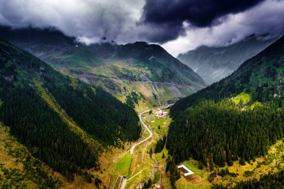 Aerial view of the Transfagarasan winding road in the Carpathian Mountains of Romania, with green slopes and cloudy skies above.