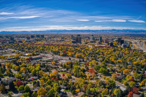 Aerial shot of Aurora, Colorado displays suburban scenery, autumn trees, streets, buildings, and distant mountains under a clear sky.