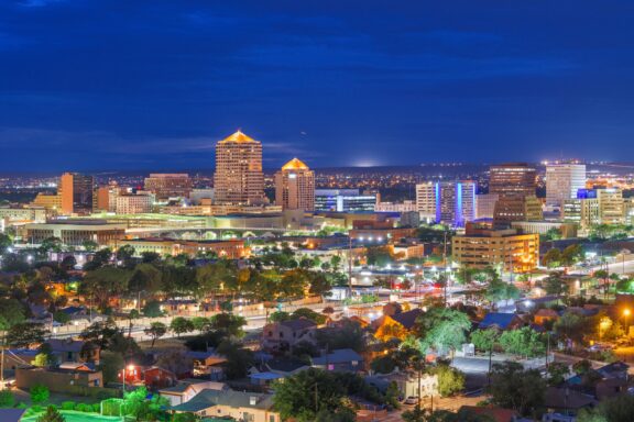 A nighttime cityscape of Albuquerque, New Mexico, featuring illuminated buildings and streets with a backdrop of a dark blue sky.