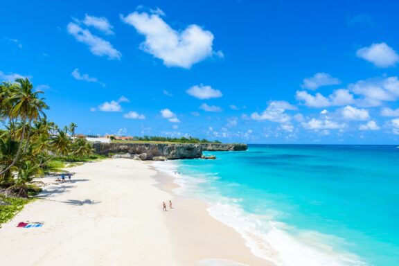 A tropical beach at Bottom Bay, Barbados with white sand, turquoise water, a few people, and lush greenery on the cliffs.