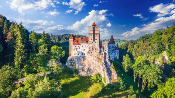 Aerial view of Bran Castle, a historic fortress on a hill surrounded by trees, under a clear sky in Romania.