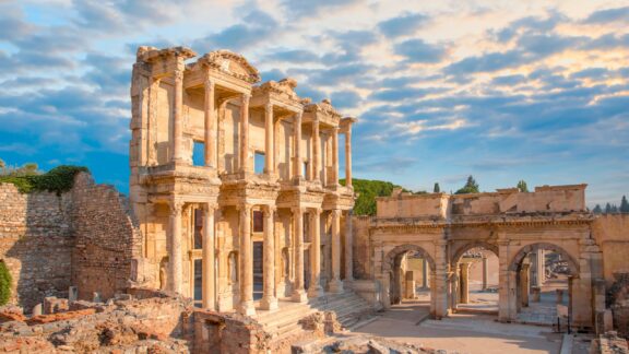 A sunrise/sunset photo of the ornate Celsus Library in Ephesus, Turkey, under a partly cloudy sky.