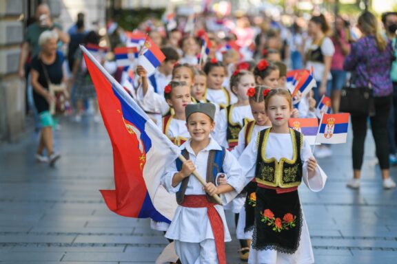 A group of children dressed in traditional Serbian folk costumes, participating in a parade, holding Serbian flags.