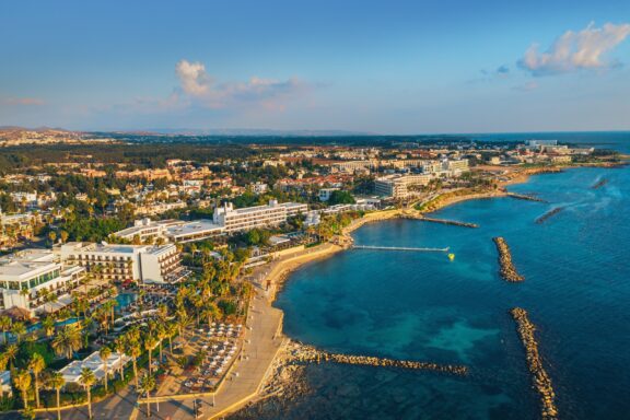 Aerial view of Paphos embankment in Cyprus displays coastline, blue waters, beaches, a jetty, and nearby buildings in daylight.