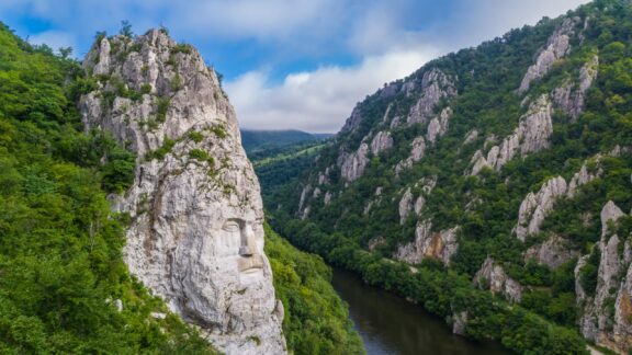 Aerial view of the Decebalus head sculpture on the rock face near the Danube River in Romania, surrounded by green forested hills.