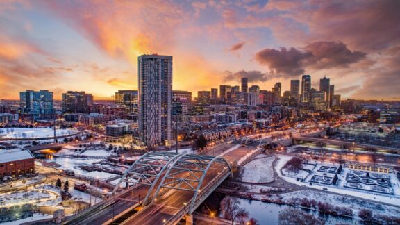 A panoramic view of downtown Denver, Colorado, USA, during sunset with snow-covered areas, prominent buildings, and a colorful sky.