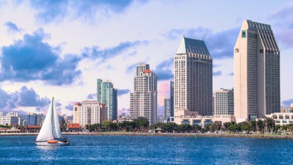 A scenic view of downtown San Diego, California, featuring skyscrapers and a sailboat on the water under a partly cloudy sky.