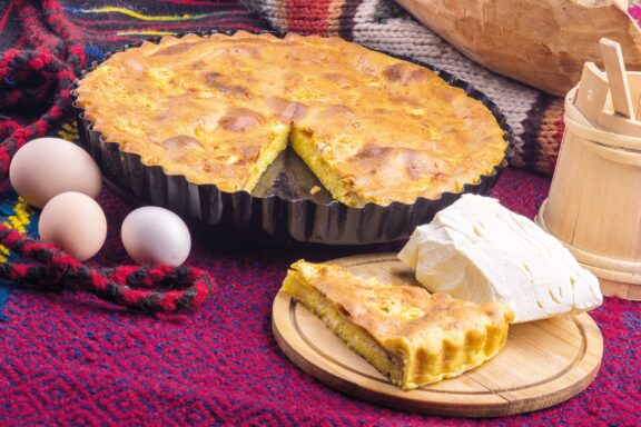 A traditional Albanian Flija pie is shown with a slice on a wooden plate, alongside fresh ingredients on a colorful cloth.