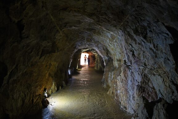 A dimly lit cave passage with rocky walls and a smooth path leading towards a light source at the end, located in Gadime Cave, Kosovo.