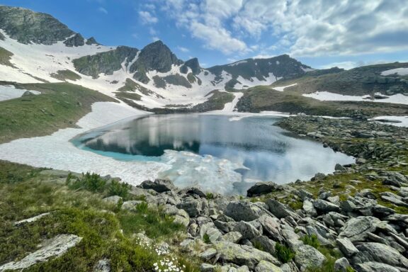 A picturesque view of Gjeravica Lake in Kosovo, with snowy mountains, a clear blue lake with ice, under a semi-cloudy sky.