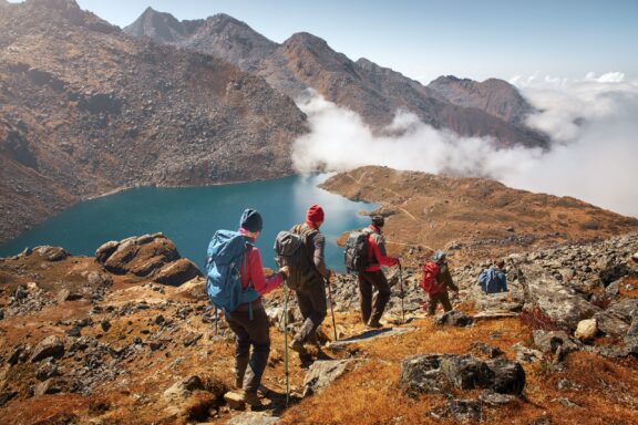 A group of tourists with backpacks hiking in the mountainous terrain of Nepal, with a clear view of a lake and clouds in the background.