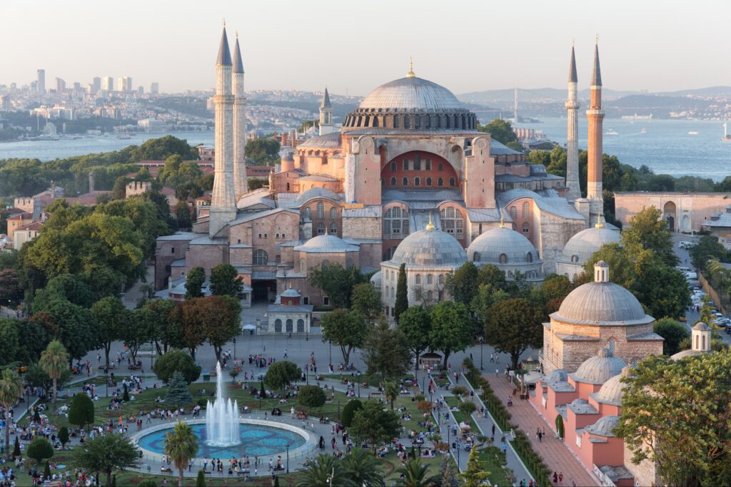 A picturesque view of Istanbul's Hagia Sophia, with its dome and minarets, framed by trees, a fountain, and cityscape backdrop.