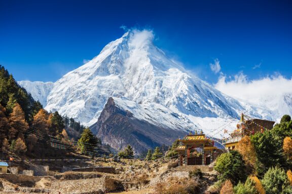 A scenic view of the snow-capped Mt. Manaslu in the Himalayas, Nepal, with a clear blue sky and a Buddhist monastery in the foreground.