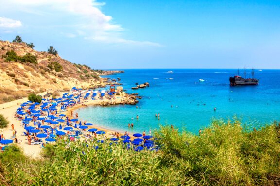 A scenic view of Konnos Beach in Cyprus, featuring clear blue waters, a sandy shore lined with blue umbrellas, and a lush green hillside.