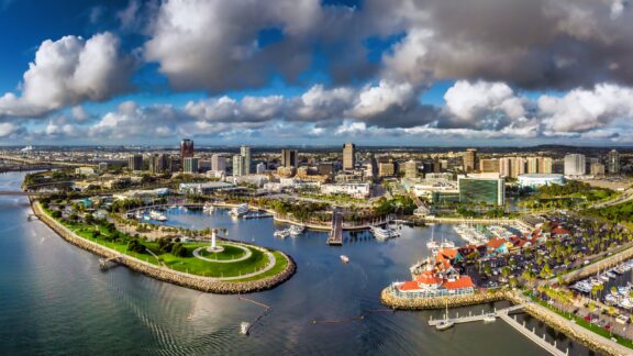 Aerial view of Long Beach, California, showcasing the coastline, lighthouse, and urban landscape under a partly cloudy sky.