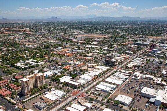 Aerial view of Main Street in downtown Mesa with buildings, streets, and vehicles, set against a backdrop of a mountain range under a clear sky.