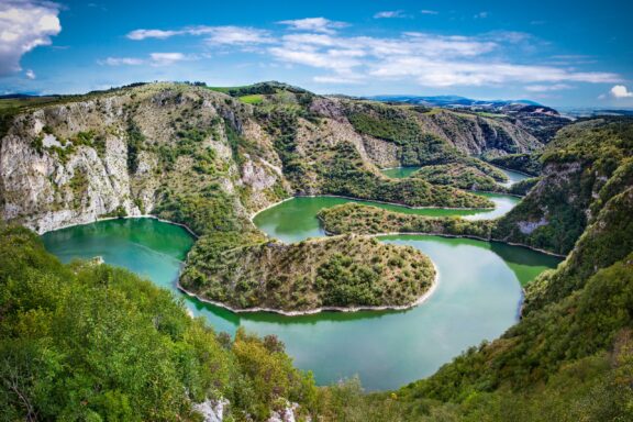 Aerial view of a meandering rocky river in Serbia, with lush greenery surrounding the winding water and hills in the background.