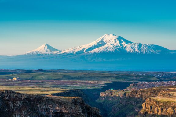 A view of Mount Ararat from Saghmosavank, Turkey, featuring the snow-capped peaks against a clear blue sky with a vast landscape in the foreground.