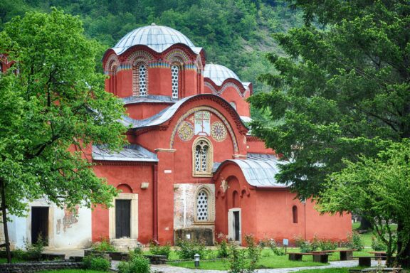 A Serbian Orthodox monastery with red walls and distinctive architectural features, nestled among lush green trees.