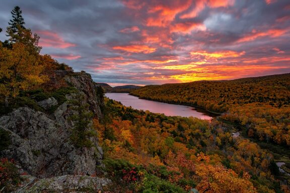 A vibrant sunset over a forested landscape with autumn foliage, reflecting on a calm river, possibly in Porcupine, Michigan.