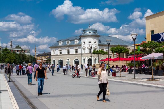 A sunny day in Pristina, Kosovo features a bustling pedestrian street, a historic building, and outdoor cafes.