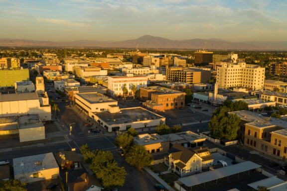 Aerial view of downtown Bakersfield with buildings, streets, and trees during golden hour, with mountains in the distance.