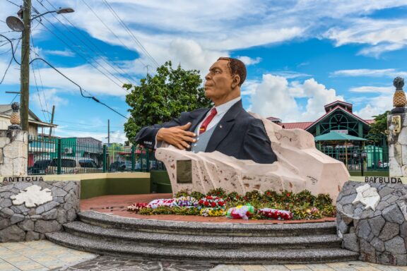 A statue of a seated man with crossed arms is located in St. John's, Antigua, with buildings and sky as backdrop.