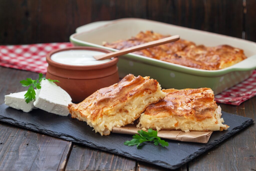 A Serbian gibanica cheese pie is served with white cheese, yogurt, and wooden chopsticks on a slate plate.