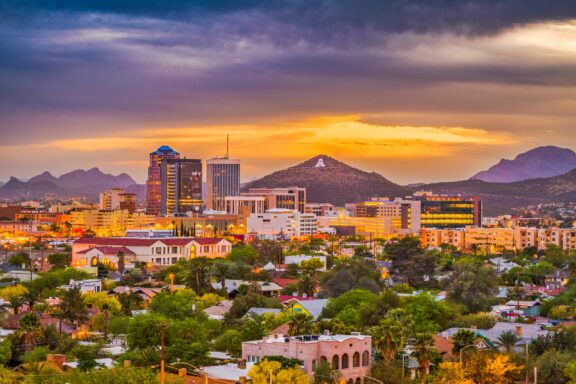 A vibrant sunset over the downtown skyline of Tucson, Arizona, with mountains in the background and scattered clouds in the sky.