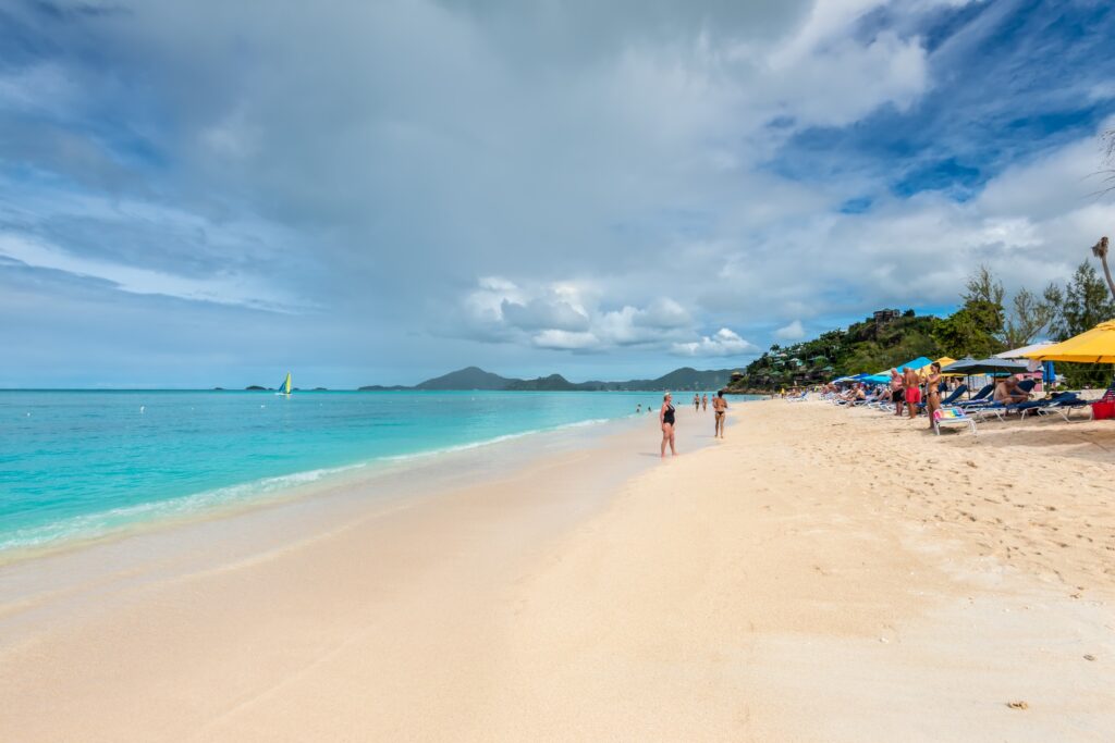 Visitors enjoy the sun at the serene sandy beach of Valley Church Bay, Antigua, with clear turquoise waters and a lush hill nearby.