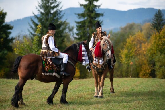 Two people in traditional Romanian attire riding horses in a field, with a backdrop of trees and hills, likely at a cultural event in Vama, Romania.