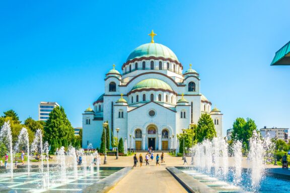 A view of Saint Sava Cathedral in Belgrade, Serbia, with fountains in the foreground and clear blue sky above.