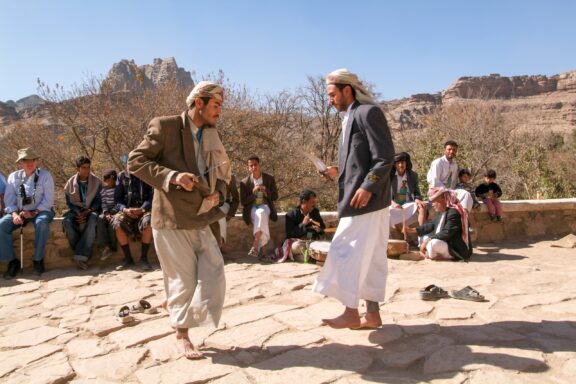 Two men in traditional clothing converse outdoors in Wadi Dhahr, Yemen, with onlookers and rocky hills nearby.