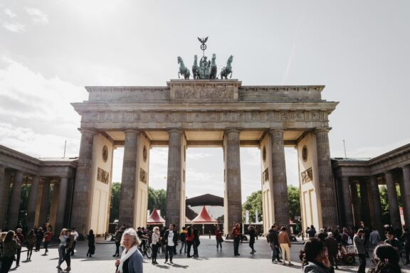 An image of the Brandenburg Gate in Germany, with people walking around in the foreground under a partly cloudy sky.