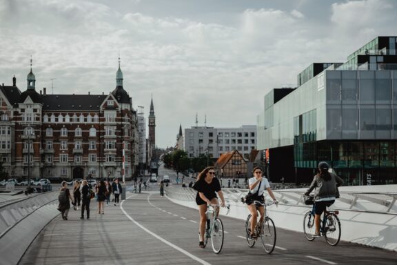 People cycle and walk on a wide pathway in Copenhagen, Denmark, with a backdrop of traditional and modern architecture.