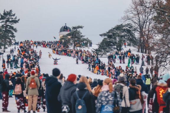 A crowd of people gathered in a snowy park in Helsinki, Finland, with some individuals taking photos and others watching an event.