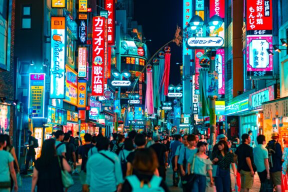 A vibrant street scene at night in Shibuya, Tokyo, Japan, with illuminated signage in various colors and a crowd of people.