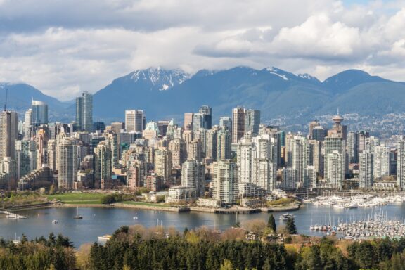 A panoramic view of Vancouver's dense cityscape, high-rises, surrounded by water and mountains under a partly cloudy sky.