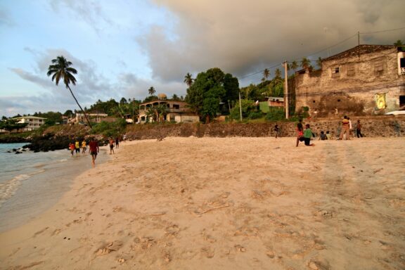 A sandy beach at sunset with people and buildings in the background, located in Moroni, Grand Comoros.