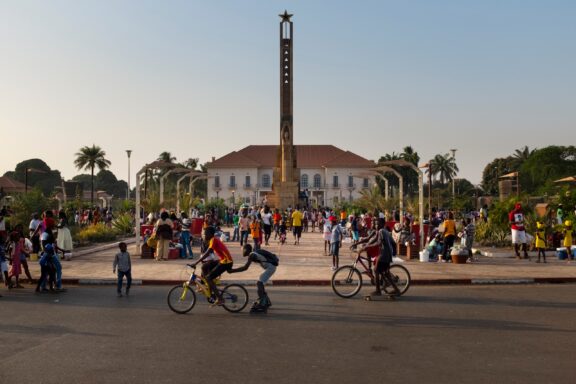 A vibrant scene in Bissau, Guinea-Bissau, featuring a public square, colonial architecture, a monument, and surrounding greenery.