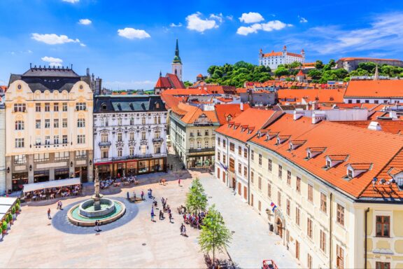 Main square in Bratislava features historical buildings, a central fountain, and distant view of Bratislava Castle under a clear sky.