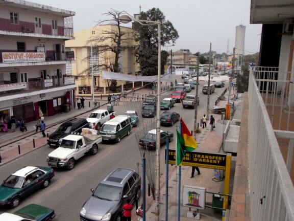 A busy Brazzaville street in Congo, Africa, with cars, buildings, pedestrians, a visible flag, and an overcast sky.