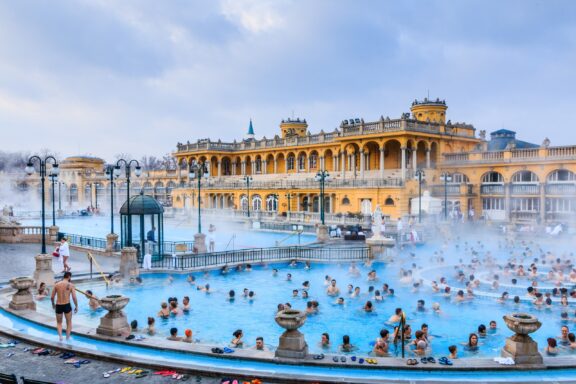 People relax in a busy outdoor thermal bath in Budapest, Hungary, with the yellow Szechenyi Bath building in the background.