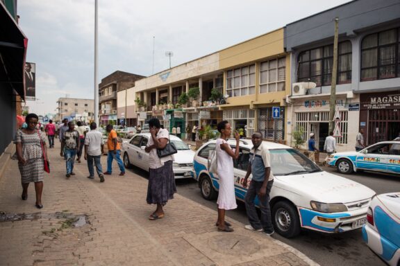 A busy street scene in Bujumbura, Burundi, with pedestrians and parked cars along the roadside, and buildings with storefronts in the background.