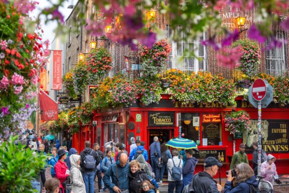 People walking on a bustling street in Dublin, Ireland, with colorful flowers adorning the buildings and a vibrant red pub in the background.