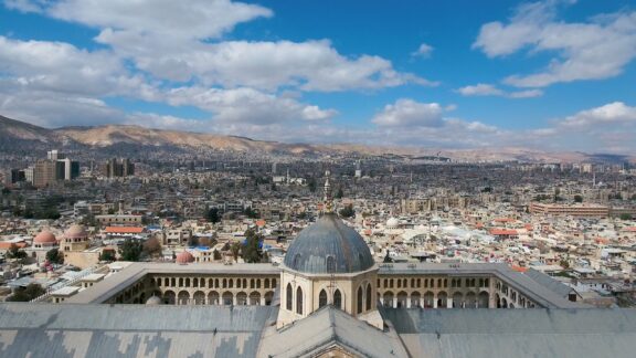 A scenic view of Damascus featuring the Great Mosque of the Umayyads, cityscape, mountains, and a partly cloudy sky.